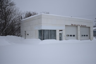 A small industrial building is surrounded by a thick layer of snow. The building has a flat roof and is made of off-white panels. Two large closed garage doors are visible, with one smaller entrance in between. Bare trees stand on the left side, and accumulation of snow is evident on the ground and building surfaces.
