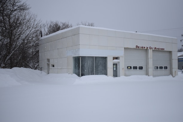 A small industrial building is surrounded by a thick layer of snow. The building has a flat roof and is made of off-white panels. Two large closed garage doors are visible, with one smaller entrance in between. Bare trees stand on the left side, and accumulation of snow is evident on the ground and building surfaces.