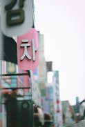 A close-up view of a pink sign with white Korean characters, hanging among other colorful signs on a street filled with various advertisements and shop signs.