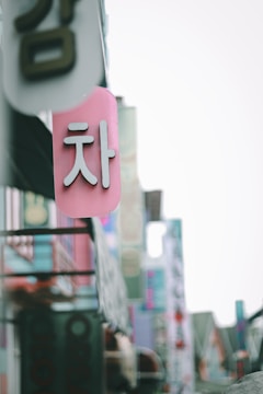 A close-up view of a pink sign with white Korean characters, hanging among other colorful signs on a street filled with various advertisements and shop signs.