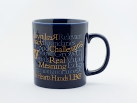 A black ceramic mug adorned with multiple motivational words and phrases in various fonts and sizes. The text is primarily in shades of white and gold, creating a striking contrast against the dark background.