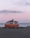 A vintage orange van with a pop-up roof is parked by the seaside. The ocean stretches out in the background, and the sky is a soft pink and purple, suggesting either sunrise or sunset. The van appears to be equipped for camping and sits on an empty paved area next to a low fence.