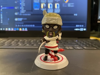 A small toy figure with a cartoonish design standing on a laptop keyboard. The figure is wearing a white outfit with red accents and a headband, holding what appears to be a lollipop. The background consists of a laptop screen displaying a software interface with various icons and text.