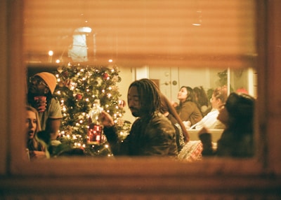 A cozy indoor party scene viewed through a window, featuring a group of people socializing around a decorated Christmas tree adorned with lights and ornaments. Participants are holding drinks and appear to be enjoying each other's company.