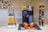 A Nespresso coffee machine is placed in the center with a stack of coffee capsules organized in a metal stand to the right. To the left, a knife block holds several knives. Orange coffee cups are positioned on a mat in front of the coffee machine. Decorative letters form the word 'cafe' on the counter, with a mug that has a 'fresh coffee' design on top.