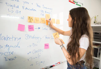 A woman with long hair is writing on a whiteboard. She is using sticky notes and markers to organize and plan a strategy. The board has various handwritten notes and symbols related to marketing and digital education.