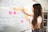 A woman with long hair is writing on a whiteboard. She is using sticky notes and markers to organize and plan a strategy. The board has various handwritten notes and symbols related to marketing and digital education.