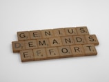 Wooden Scrabble tiles arranged to spell the phrase 'GENIUS DEMANDS EFFORT', placed on a light surface.