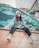 A person is sitting on the edge of a tiled wall, chewing bubble gum. They are wearing a casual outfit with a gray beanie, a gray sweatshirt, and black pants, complemented by white sneakers. The background features a wall with a colorful mural depicting large leaves and flowers.