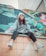 A person is sitting on the edge of a tiled wall, chewing bubble gum. They are wearing a casual outfit with a gray beanie, a gray sweatshirt, and black pants, complemented by white sneakers. The background features a wall with a colorful mural depicting large leaves and flowers.