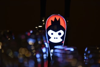 A beer tap handle featuring a stylized, illuminated monkey face with a crown design against a red and black background. The surrounding area has an out-of-focus effect, highlighting bokeh lights and blurred glassware, creating a dimly lit atmosphere.