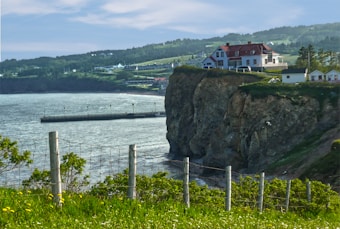 A coastal scene depicts a large house perched on a cliff overlooking the ocean. The house is surrounded by lush greenery and a fence runs along the cliff edge. The sea extends into the distance, bordered by a hilly landscape with more greenery.