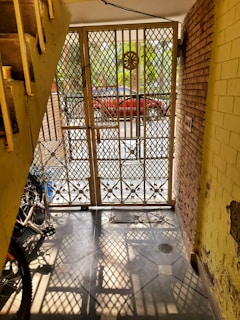 A wrought iron gate with intricate patterns stands open, allowing sunlight to cast geometric shadows on the tiled floor. A bicycle is partially visible near the stairs on the left, while a red car is parked outside against a backdrop of green foliage. The setting includes brick and yellow painted walls with some peeling sections.