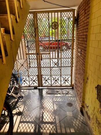 A wrought iron gate with intricate patterns stands open, allowing sunlight to cast geometric shadows on the tiled floor. A bicycle is partially visible near the stairs on the left, while a red car is parked outside against a backdrop of green foliage. The setting includes brick and yellow painted walls with some peeling sections.
