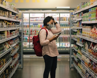 A person wearing a mask stands in the aisle of a grocery store, holding a phone and appearing to browse or compare products on the shelves. The scene includes a variety of packaged goods, with colorful items lining both sides of the aisle.
