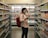 A person wearing a mask stands in the aisle of a grocery store, holding a phone and appearing to browse or compare products on the shelves. The scene includes a variety of packaged goods, with colorful items lining both sides of the aisle.