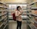 A person wearing a mask stands in the aisle of a grocery store, holding a phone and appearing to browse or compare products on the shelves. The scene includes a variety of packaged goods, with colorful items lining both sides of the aisle.