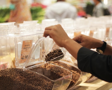 Hands are scooping coffee beans from a transparent container in a store setting. The container is labeled with a price and product name. The background is blurred, indicating a busy retail environment.