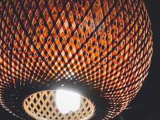 A close-up view of a woven rattan lampshade with an intricate lattice pattern, illuminated from the inside by a bright light bulb. The pattern creates a play of light and shadow, highlighting the warm, earthy tones of the material against a dark background.