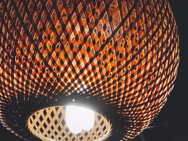 A close-up view of a woven rattan lampshade with an intricate lattice pattern, illuminated from the inside by a bright light bulb. The pattern creates a play of light and shadow, highlighting the warm, earthy tones of the material against a dark background.