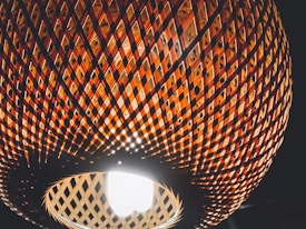 A close-up view of a woven rattan lampshade with an intricate lattice pattern, illuminated from the inside by a bright light bulb. The pattern creates a play of light and shadow, highlighting the warm, earthy tones of the material against a dark background.