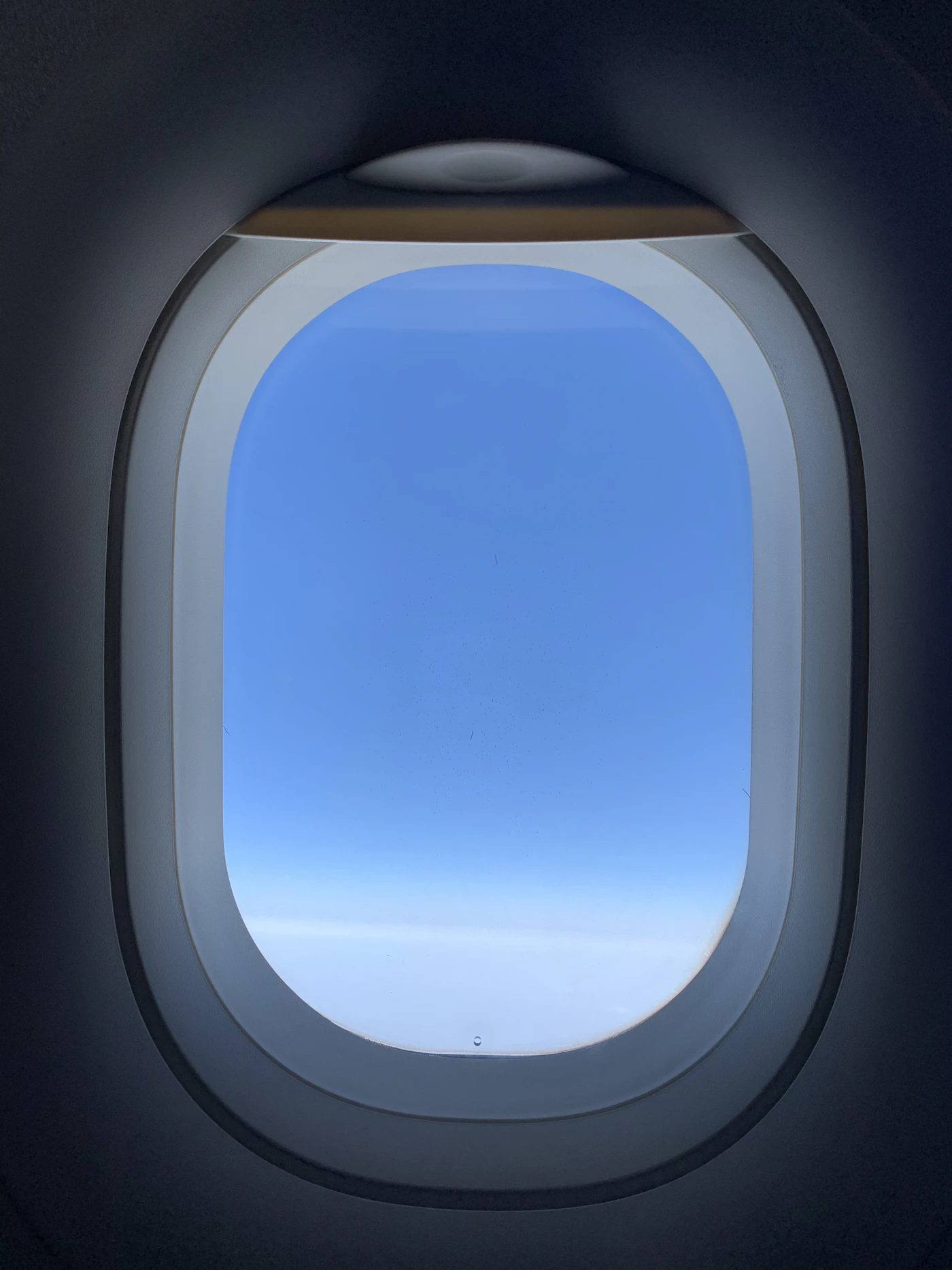 A calm view out a cabin window of clear blue sky above a soft layer of white clouds during a smooth cruise