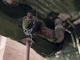 A rusted pipe under a structure is covered with green moss, with water dripping from a joint. The surrounding area shows signs of decay, with cracked surfaces and discolored materials.