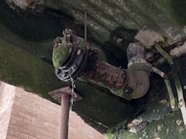 A rusted pipe under a structure is covered with green moss, with water dripping from a joint. The surrounding area shows signs of decay, with cracked surfaces and discolored materials.