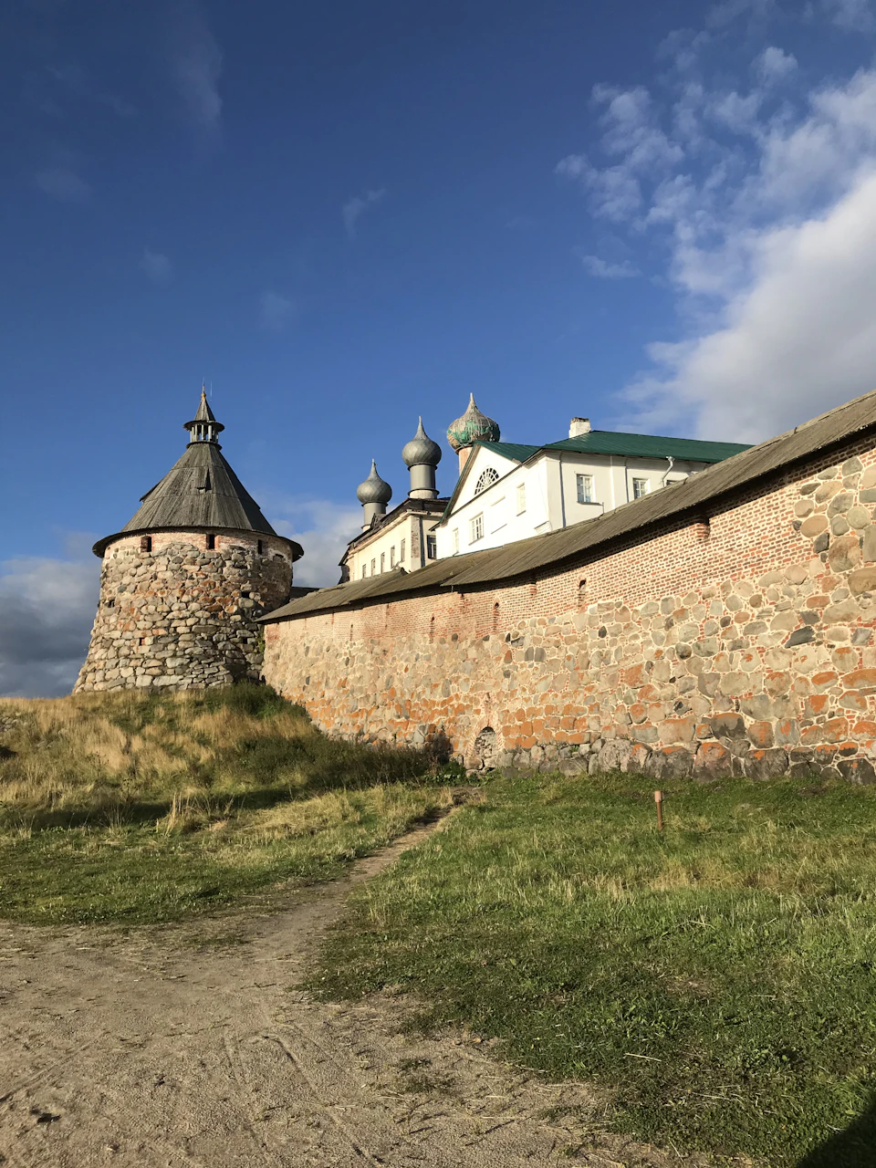 The image depicts a stone wall with a tall tower on top, situated within an open field under a clear blue sky. The perspective is from ground level, looking up at the imposing structure and the vast expanse of the surrounding landscape. In the distance, beyond the stone wall, there are buildings visible in the background, suggesting that this location may be part of a larger city or town.

The image does not contain any discernible text or countable objects, but it is clear that the scene captures the essence of an open field with a historical and architectural element.