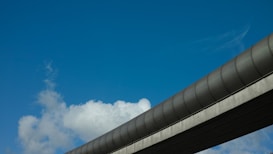 A blue sky serves as the backdrop for a modern, elevated structure resembling a bridge or monorail track. White clouds are scattered across the sky, adding depth to the scene.