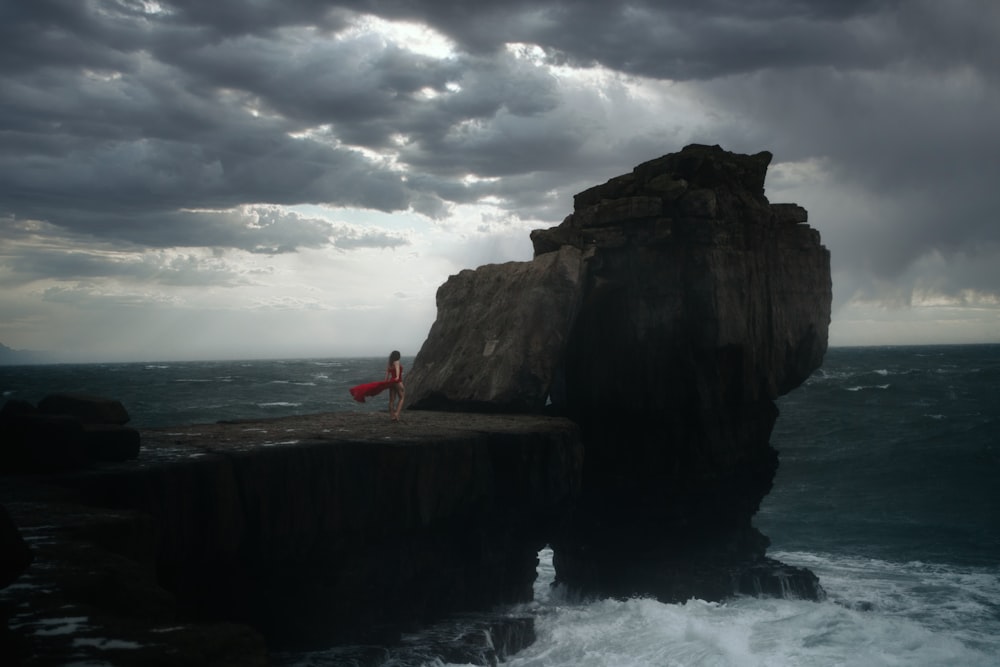 person in red shirt standing on rock formation near sea under cloudy sky during daytime
