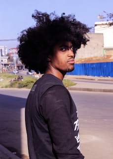 A person with a large afro hairstyle stands on an urban street, looking over their shoulder. The background includes a cityscape with buildings and a blue fence, suggesting an outdoor setting under bright sunlight.