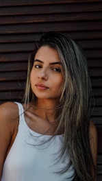 A woman with long, dark hair is gazing directly at the camera. She is wearing a white sleeveless top. The background consists of horizontal wooden planks.