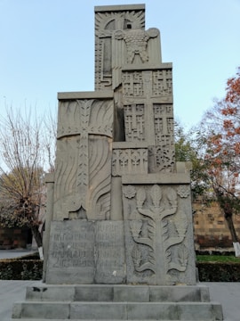 A stone monument with intricate carvings, including decorative patterns, crosses, and what appears to be text in a script. The sculpture features motifs of leaves and an ornate relief of a ram. Trees and a stone wall can be seen in the background, under a clear blue sky.