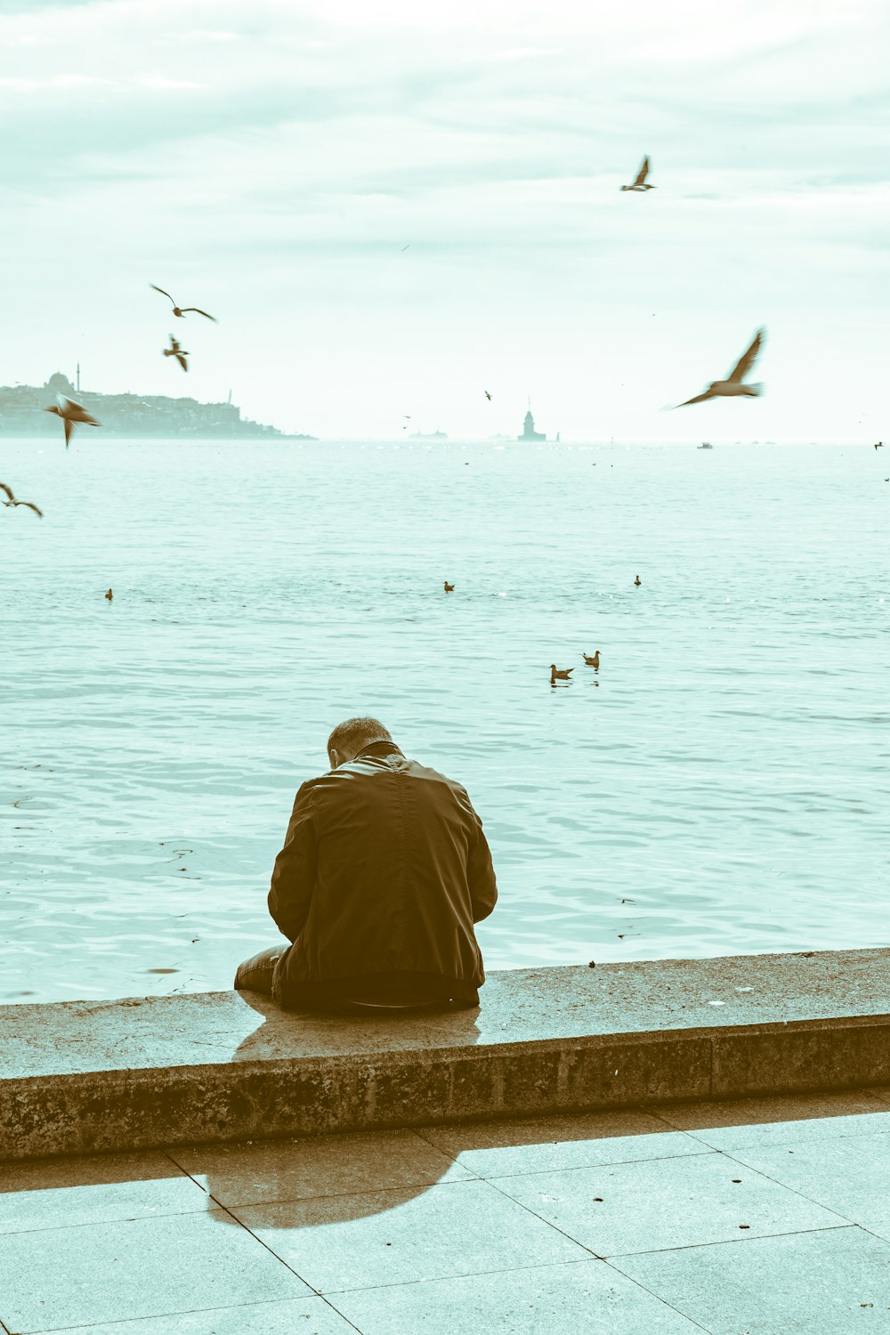 man in brown hoodie sitting on concrete bench looking at birds flying over the sea during