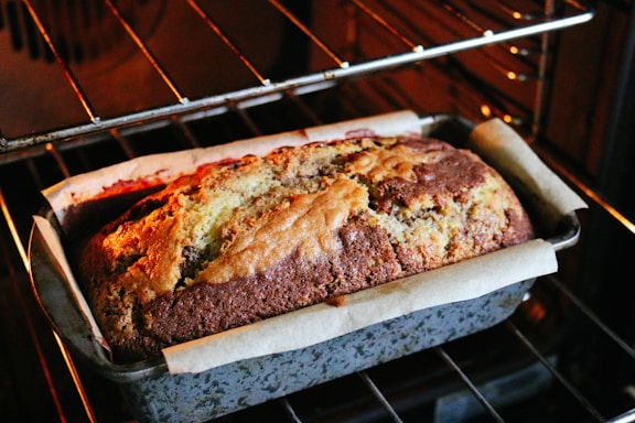 A loaf of cake with a golden brown crust, partially rising in an oven. The cake is set in a lined baking tin, showcasing swirls of chocolate marbling.