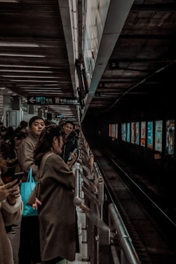 A group of people stands on a subway platform, some looking at their phones while others look down the track. Overhead signage indicates lines available from the platform. The lighting is dim, creating a moody, urban atmosphere, with the platform extending into darkness.