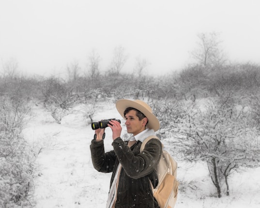 A person wearing a wide-brimmed hat and an outdoor jacket is holding binoculars, standing in a snowy landscape. The background features snow-covered shrubs and leafless trees under a gray, overcast sky. The individual appears to be intently observing something in the distance.