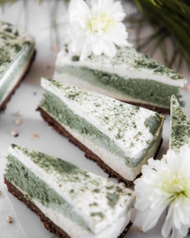 Three slices of cake with a green and white layered appearance are elegantly arranged on a surface. The cakes have a dusting of green powder on top and are placed alongside delicate white flowers, adding a touch of freshness and decoration.