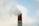 A tall, red brick chimney emits a thick cloud of smoke against a cloudy blue sky. The structure shows signs of weathering, with some areas of the brickwork appearing slightly worn.