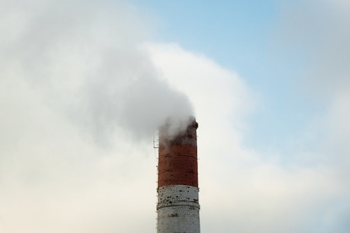 A tall, red brick chimney emits a thick cloud of smoke against a cloudy blue sky. The structure shows signs of weathering, with some areas of the brickwork appearing slightly worn.