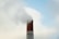 A tall, red brick chimney emits a thick cloud of smoke against a cloudy blue sky. The structure shows signs of weathering, with some areas of the brickwork appearing slightly worn.