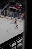 A hockey player in a red uniform is on the ice moving towards the goal, preparing to take a shot. Another player and a goalie are nearby, also in red uniforms. The arena is sparsely filled with spectators, and a large black balloon with a red star logo is visible above the stands.