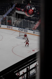 A hockey player in a red uniform is on the ice moving towards the goal, preparing to take a shot. Another player and a goalie are nearby, also in red uniforms. The arena is sparsely filled with spectators, and a large black balloon with a red star logo is visible above the stands.