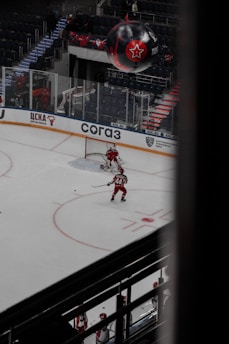 A hockey player in a red uniform is on the ice moving towards the goal, preparing to take a shot. Another player and a goalie are nearby, also in red uniforms. The arena is sparsely filled with spectators, and a large black balloon with a red star logo is visible above the stands.