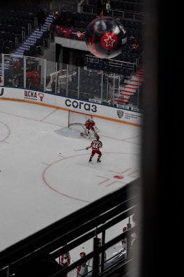 A hockey player in a red uniform is on the ice moving towards the goal, preparing to take a shot. Another player and a goalie are nearby, also in red uniforms. The arena is sparsely filled with spectators, and a large black balloon with a red star logo is visible above the stands.