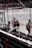 Hockey players in red and white uniforms are exiting the ice rink through a gate. The image captures the back view of these players as they move towards a bench area. Spectators are visible in the stands, and banners with advertisements are displayed around the rink.