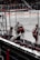 Hockey players in red and white uniforms are exiting the ice rink through a gate. The image captures the back view of these players as they move towards a bench area. Spectators are visible in the stands, and banners with advertisements are displayed around the rink.