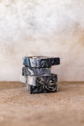 A stack of artisanal soap bars with a marbled design in shades of black, gray, and white, placed on a textured surface against a blurred neutral background.