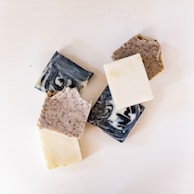 A collection of various handmade soap bars with different textures and colors, including marbled black and white, speckled, and plain white soaps, arranged on a white surface.